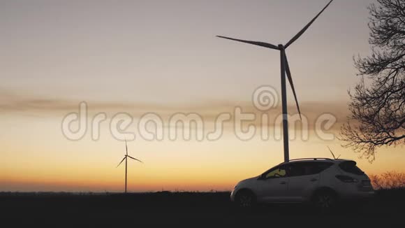 日落时分风力涡轮机背景下的白色汽车视频的预览图