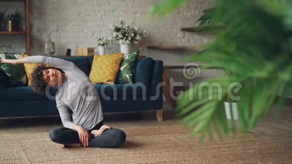 长得好看的卷发女孩伸着胳膊扭着身子坐在地毯上轻松的莲花姿势视频的预览图