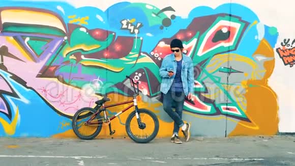 一个骑自行车的人站在墙边在他的手机上打字年轻时尚的白人男性青少年视频的预览图