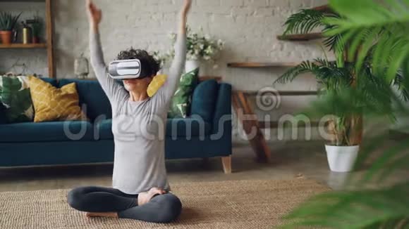 戴着虚拟现实眼镜的瘦女孩坐在家里的地板上坐在莲花的位置上享受着个人的乐趣视频的预览图