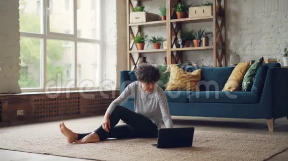 年轻漂亮的女士正从网上学习瑜伽坐在地上看手提电脑屏幕练习身体视频的预览图