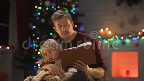 专心致志的爸爸在装饰好的树旁为困倦的女孩读圣诞童话视频的预览图
