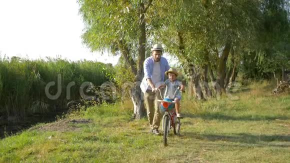 小男孩学着骑自行车在她父亲的帮助下在农村的湖边视频的预览图