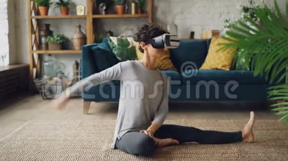 戴着虚拟现实眼镜的微笑女孩正在家中训练坐在地板上伸展腿部和身体享受AR视频的预览图