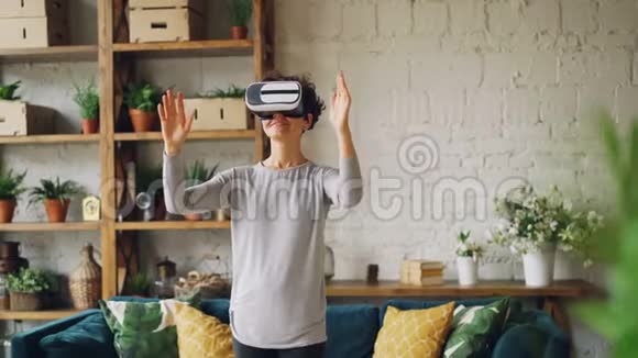 快乐女孩正在家中使用虚拟现实眼镜站在房间里双手移动微笑着玩现代小玩意视频的预览图