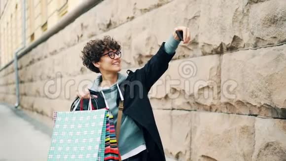 卷发女子慢动作自拍带着五颜六色的购物袋站在户外拿着智能手机和视频的预览图