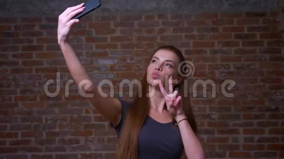 可爱的白种人女性正在自拍并在她的手机上显示她的两个手指同时站在砖背景上视频的预览图