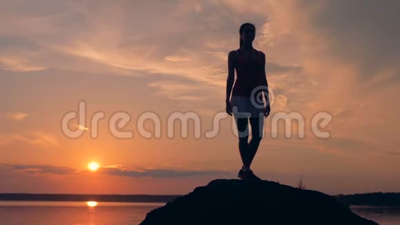 在日落时苗条的年轻女士正在湖岸上采取一种姿势慢动作视频的预览图