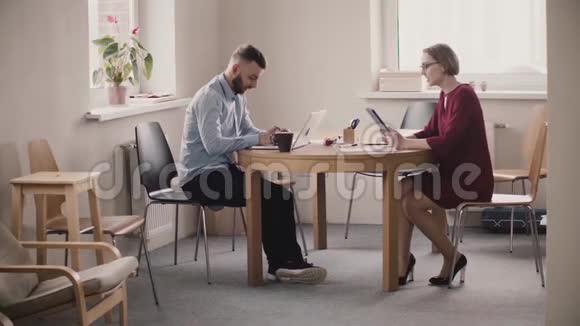 年轻的女老板坐在桌子旁边的男员工与笔记本电脑显示他的信息平板电脑在现代轻型办公室视频的预览图