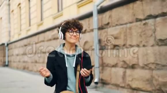 戴着时髦眼镜的漂亮黑发女郎正在耳机里听收音机在走路时使用智能手机视频的预览图