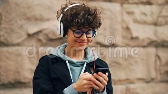 漂亮的女孩是耳机眼镜是听音乐和触摸智能手机屏幕选择歌曲站着视频的预览图