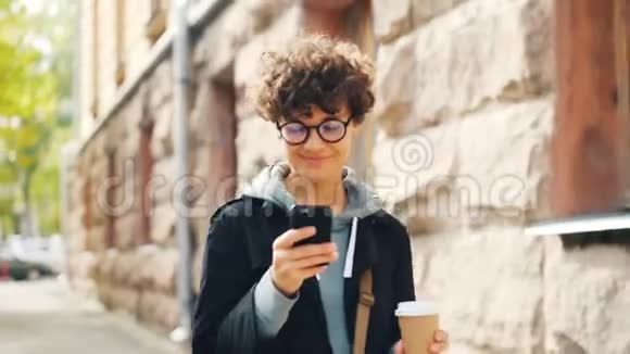 戴着眼镜的年轻女士微笑着用智能手机看屏幕一边在城市户外边走边喝咖啡视频的预览图