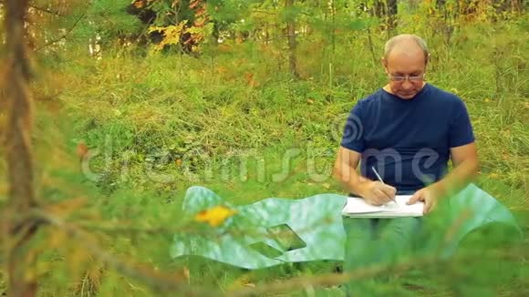 一个戴眼镜的人在森林的边缘用铅笔在笔记本上画草图视频的预览图