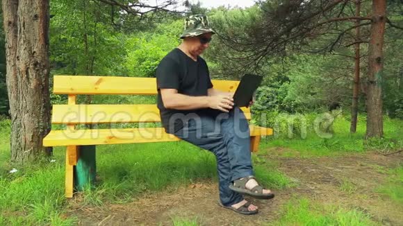 公园里一个戴太阳镜的人坐在长凳上拿着笔记本电脑工作视频的预览图
