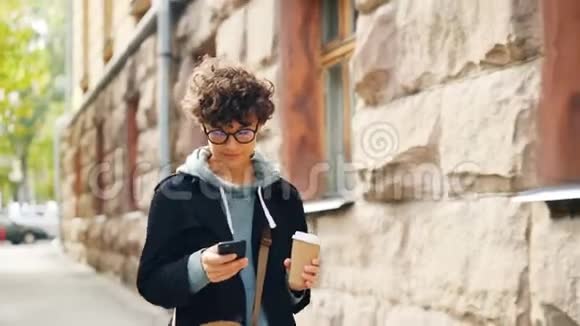 美丽的女孩深色卷发正在使用智能手机发短信给朋友并抱着咖啡独自在城市散步现代视频的预览图
