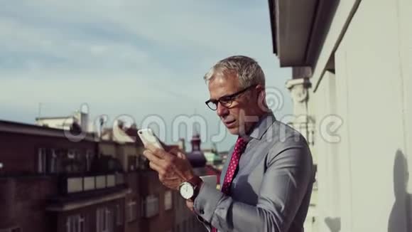 成熟的商人智能手机站在城市的露台上打电话视频的预览图