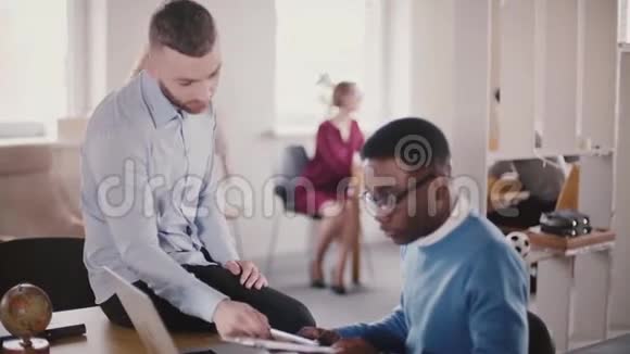 自信的年轻男性首席执行官商人坐在办公室的桌子上帮助他的非裔美国同事合作视频的预览图