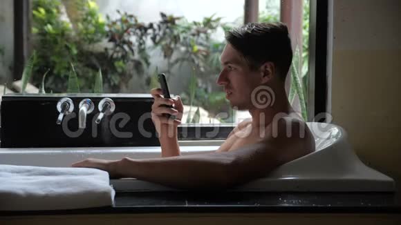 年轻英俊的男人躺在浴缸里用智能手机成功地交易微笑男人在智能手机上获得胜利信息视频的预览图