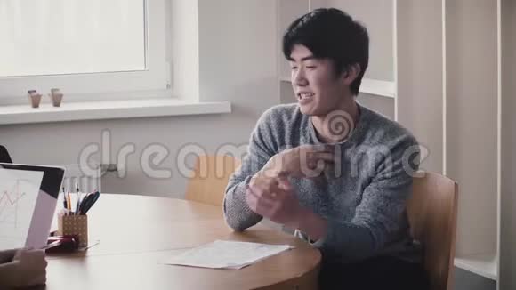年轻英俊快乐的日本男人在桌子旁说话微笑着在现代时尚的轻型办公室接受面试视频的预览图