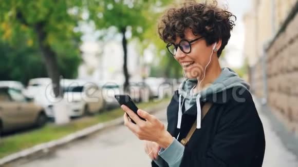 一位留着卷发和眼镜的年轻女子站在户外和朋友聊天还使用智能手机和手机视频的预览图