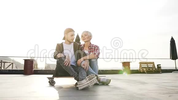 两个年轻人坐在工业大厦屋顶上的滑板上年轻夫妇坐在那里玩得开心视频的预览图