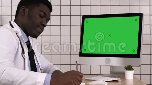 年轻的非洲医生穿着实验室外套坐在他的办公室里一边看电脑屏幕一边做一些笔记白色视频的预览图