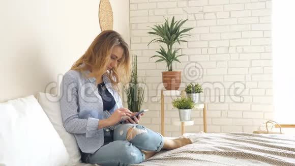 穿牛仔裤的金发女孩坐在床上做智能手机视频的预览图