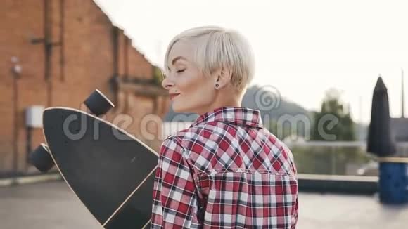 后景美丽的金发女孩与长滑板走在现代建筑屋顶美丽的年轻女子视频的预览图