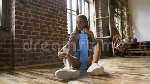 迷人的少女正坐在地板上手里拿着滑板在室内的智能手机上制作自拍照片视频的预览图