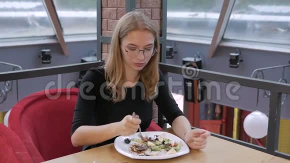 一个戴眼镜的年轻女孩坐在咖啡馆里吃着凯撒沙拉把肩膀移到音乐的节拍上视频的预览图