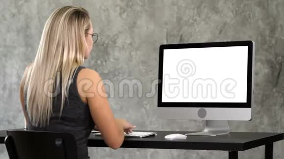 穿着时髦眼镜的嬉皮士女孩坐在电脑前的桌子上工作白色显示器视频的预览图