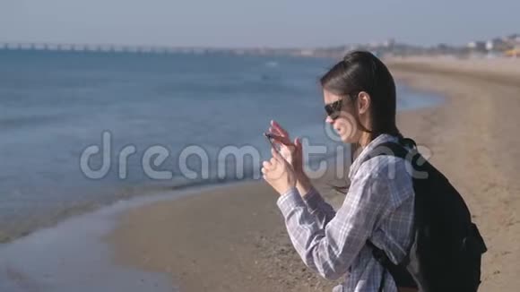旅行者女孩和一个背包博主在沙滩上并在手机上制作照片和视频视频的预览图