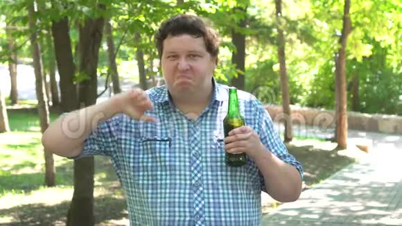 严肃的年轻胖子穿着衬衫拇指朝下站着喝着一瓶冰镇啤酒视频的预览图