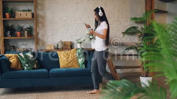 迷人的亚洲女士正在耳机里听音乐使用智能手机在客厅的地板上跳舞视频的预览图