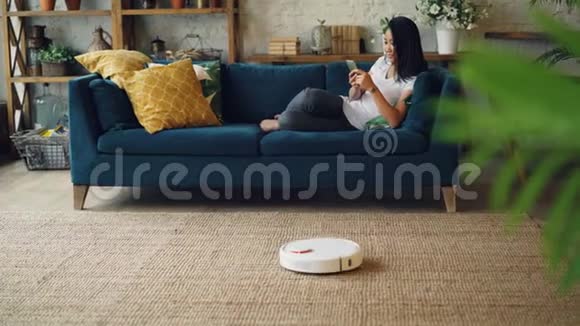 亚洲学生正在使用智能手机休息在沙发上当机器人胡佛是在地板和地毯的房子电气视频的预览图