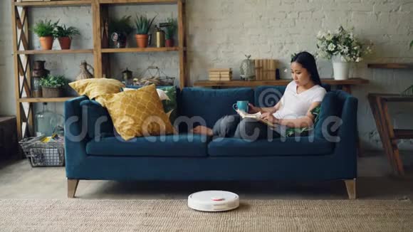 美丽的亚洲女孩正在看书躺在客厅的沙发上而机器人吸尘器则在干洗地板上视频的预览图