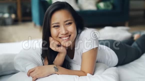 一幅美丽的亚洲女士躺在家里的床上微笑着然后笑着看着镜头放松心情好视频的预览图