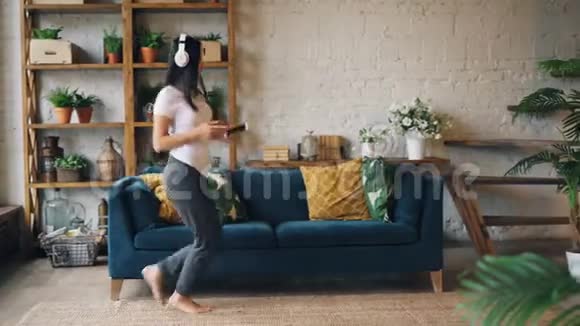 快乐的亚洲女孩正在家中的地板上跳舞用智能手机在耳机里旋转和跳跃听音乐视频的预览图