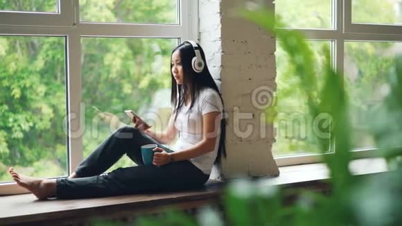 漂亮的亚洲学生正在享受音乐耳机坐在窗台上使用智能手机触摸屏幕现代视频的预览图