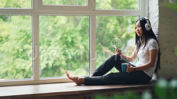 放松的亚洲女士坐在窗台上通过无线耳机听音乐使用智能手机视频的预览图