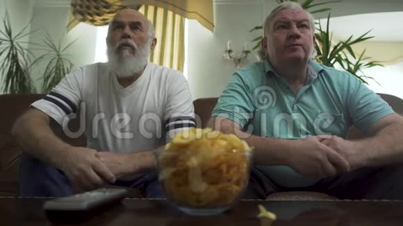 两个成熟的高级男人坐在棕色的皮革沙发上看电视退休人员的休闲视频的预览图