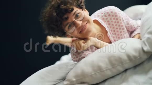戴着眼镜穿着休闲服装带着幸福的微笑和神情在床上翻滚的漂亮女士的肖像视频的预览图