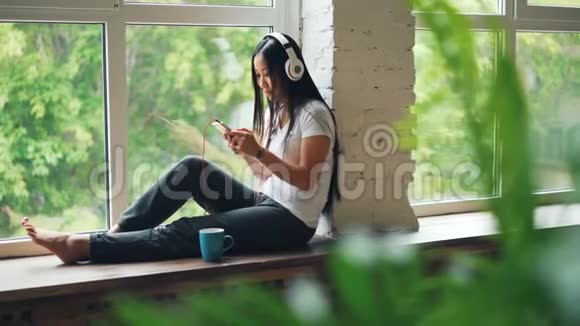 开朗的亚洲女人正在听着耳机里最喜欢的歌还在用智能手机笑着坐着视频的预览图
