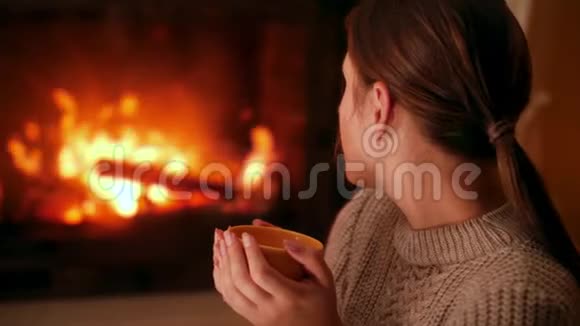年轻女子喝热茶晚上在家里壁炉里烧火取暖视频的预览图