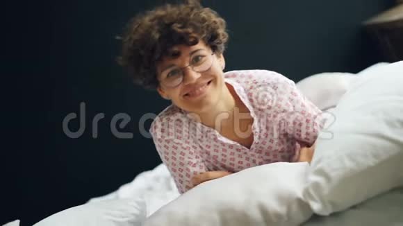 快乐女孩的肖像短卷发戴着眼镜在床上滚动微笑着看着镜头表达视频的预览图