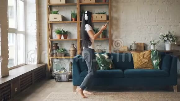 漂亮的亚洲女人独自在家跳舞通过耳机和智能手机听音乐视频的预览图