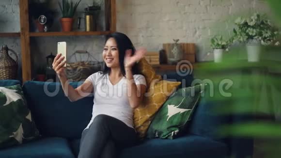 现代亚洲女孩正在制作一个在线视频打电话挥手然后在家里坐在沙发上打手势视频的预览图