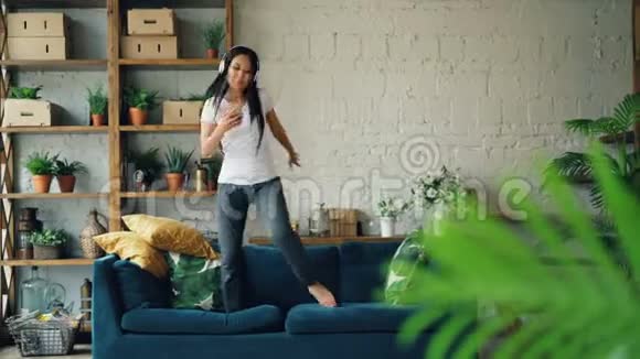 可爱的亚洲女孩正在沙发上跳舞通过耳机听智能手机音乐并在室内签名欣赏歌曲视频的预览图