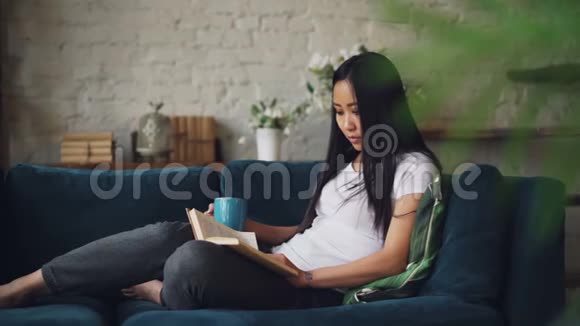 聪明的黑发年轻女士正在看书拿着一杯咖啡坐在客厅的沙发上视频的预览图
