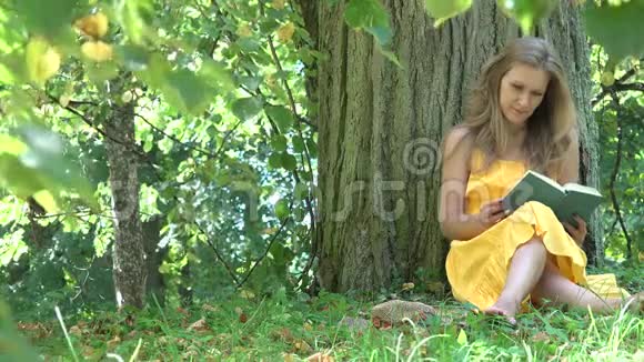 可爱的女孩子穿着黄色的衣服坐在老树下在夏天的公园里读小说4K视频的预览图